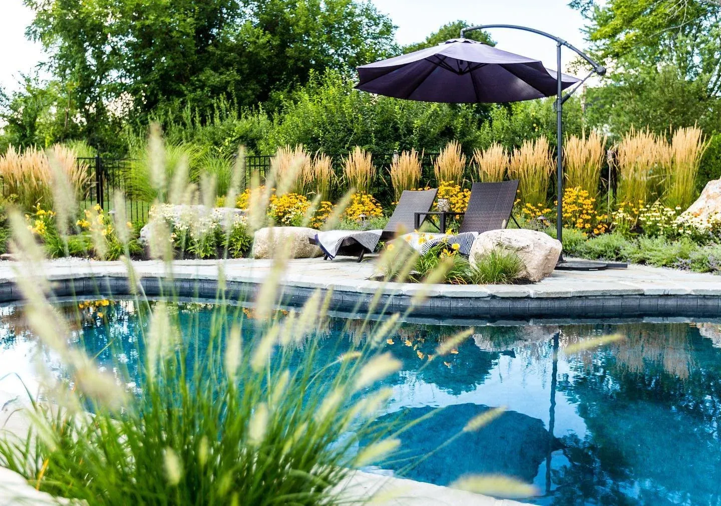 Backyard swimming pool with two lounge chairs, an umbrella, and lush greenery and flowers in the background.