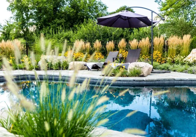 Backyard swimming pool with two lounge chairs, an umbrella, and lush greenery and flowers in the background.