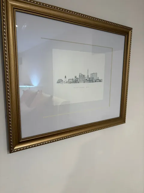 Gold-framed art piece on a white wall. The artwork is a simple, black and white sketch of a city skyline.