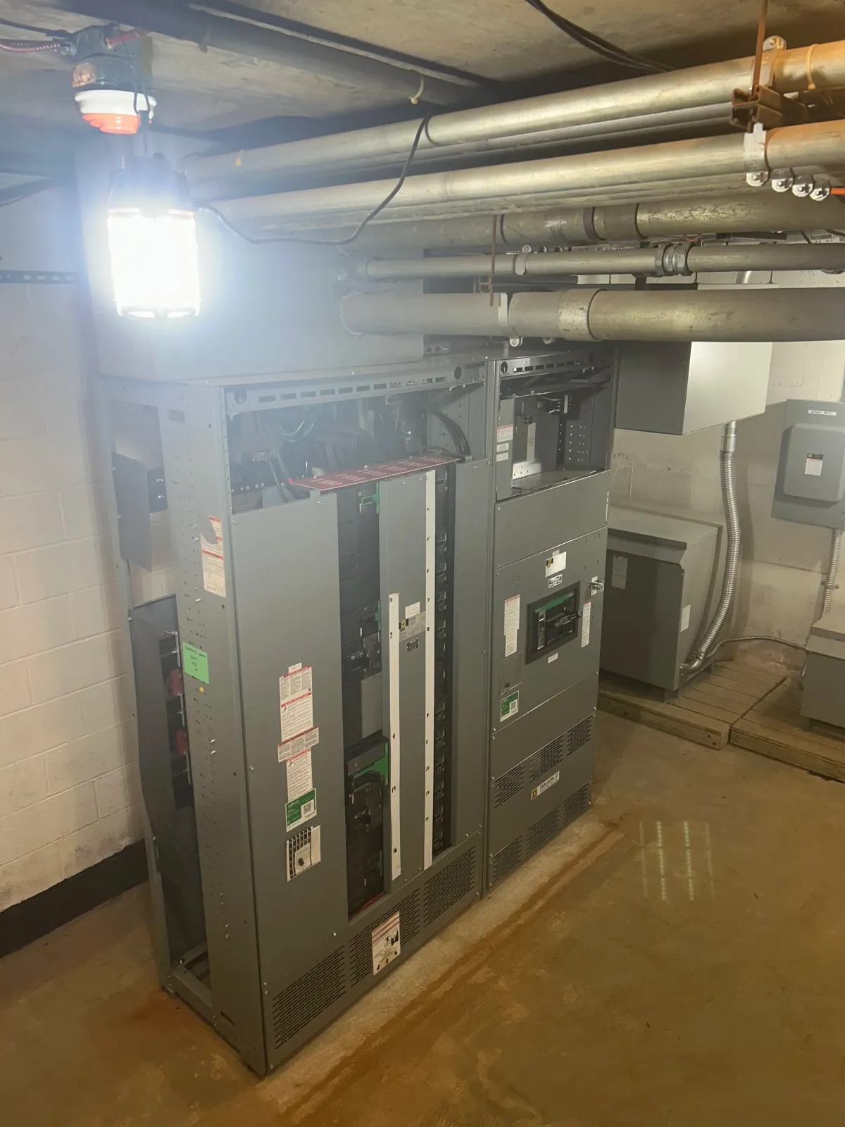 Electrical equipment and piping in a basement or utility room, with a fluorescent light overhead.
