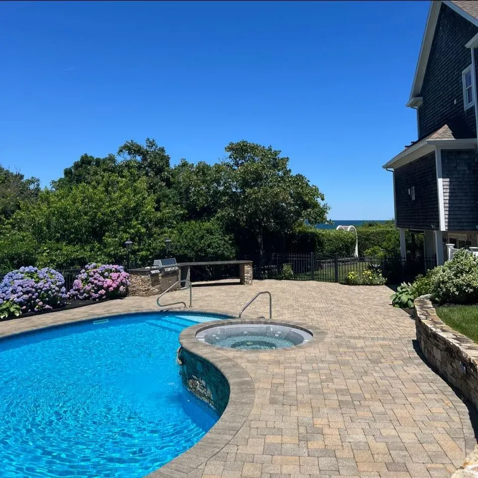Pool and spa on a brick patio with a house, landscaping, and ocean view on a sunny day.