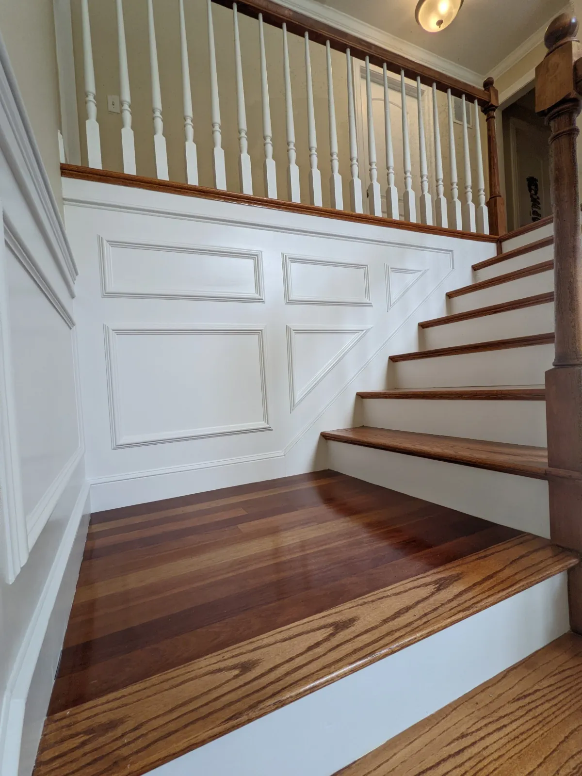 Interior view of a staircase with wooden steps and white paneled walls, featuring a white railing with spindles and a wooden handrail.