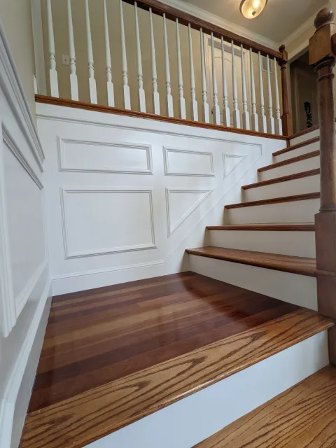 Interior view of a staircase with wooden steps and white paneled walls, featuring a white railing with spindles and a wooden handrail.
