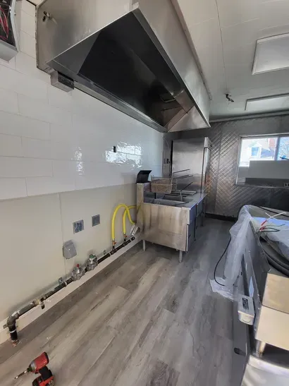 Empty commercial kitchen with stainless steel hood, white tile walls, and gray wood-look flooring.