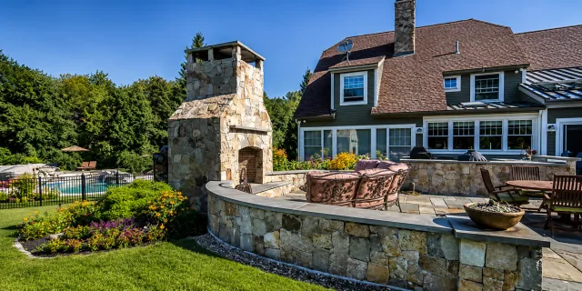 Backyard with stone outdoor fireplace, patio furniture, swimming pool, and garden, adjacent to a house with large windows and a brown shingle roof, surrounded by green trees.
