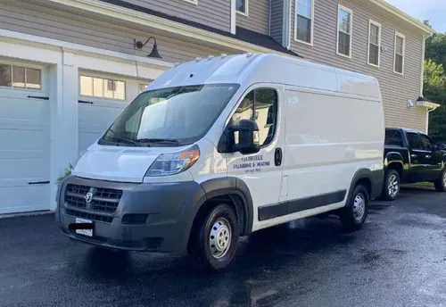 A white Ram ProMaster cargo van parked on an asphalt driveway in front of a house with a garage.