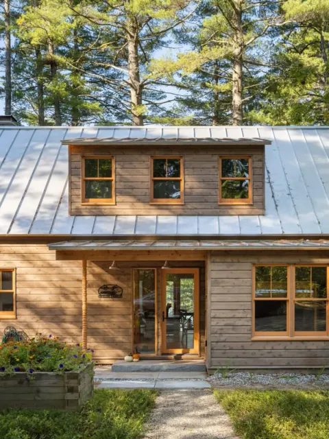 A wooden cabin with a metal roof, large windows, and a small garden bed in front, crafted by Maine homebuilders and surrounded by tall pine trees.