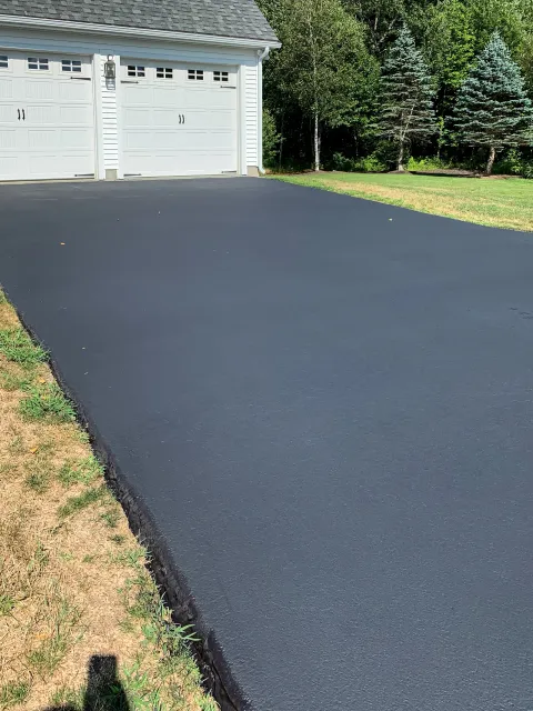 Freshly paved black asphalt driveway in front of a white garage with trees and grass on the sides.