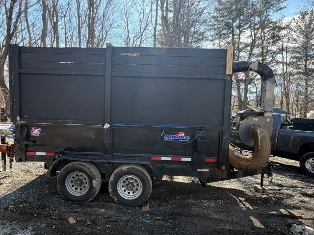 Black dump trailer with large rear chute parked outdoors beside a pickup truck in a wooded area.