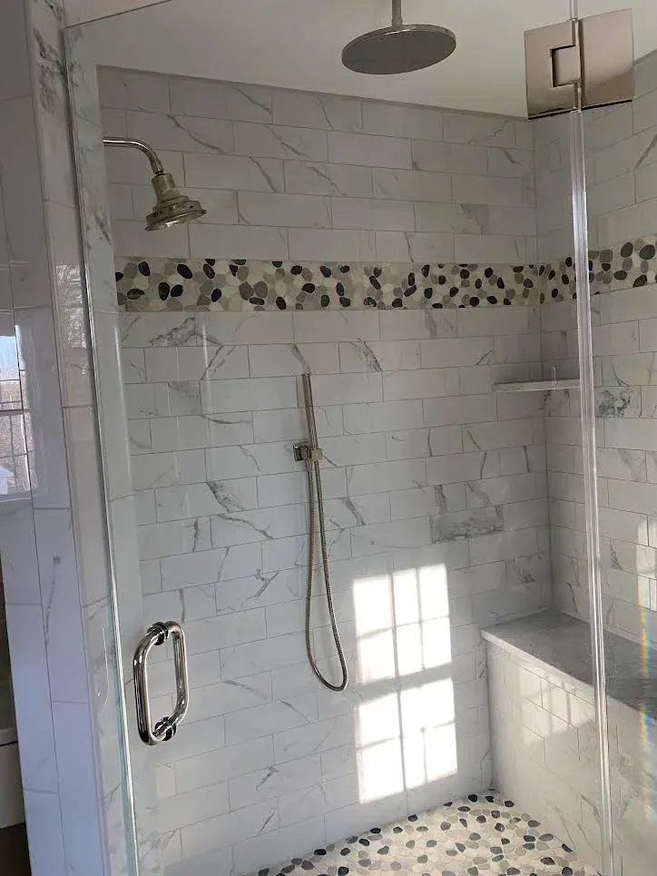 View of a modern walk-in shower with glass door, tiled walls in white marble pattern with a decorative strip of small, dark, pebble-like tiles, a rainfall showerhead, a handheld shower, a built-in ledge, and marble flooring with pebble accents.