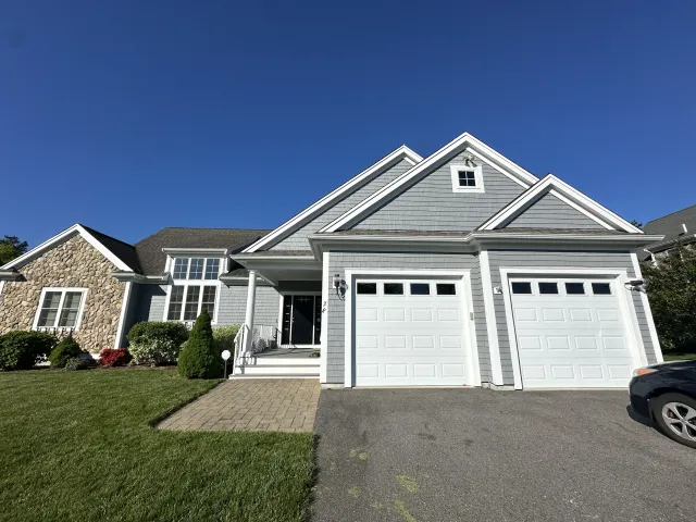 Front view of a two-story house with gray siding, white trim, a double garage, and a paved driveway on a bright, clear day.
