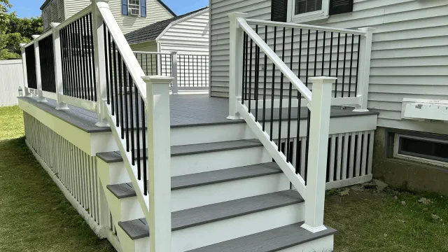 New gray wooden porch stairs with black metal railing and white support posts leading up to a house with white siding.