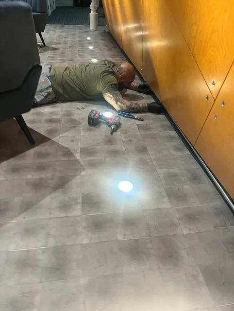 Electrician Working on the Floor, Installing or Fixing a Wooden Panel with a Drill in a Well-Lit Room