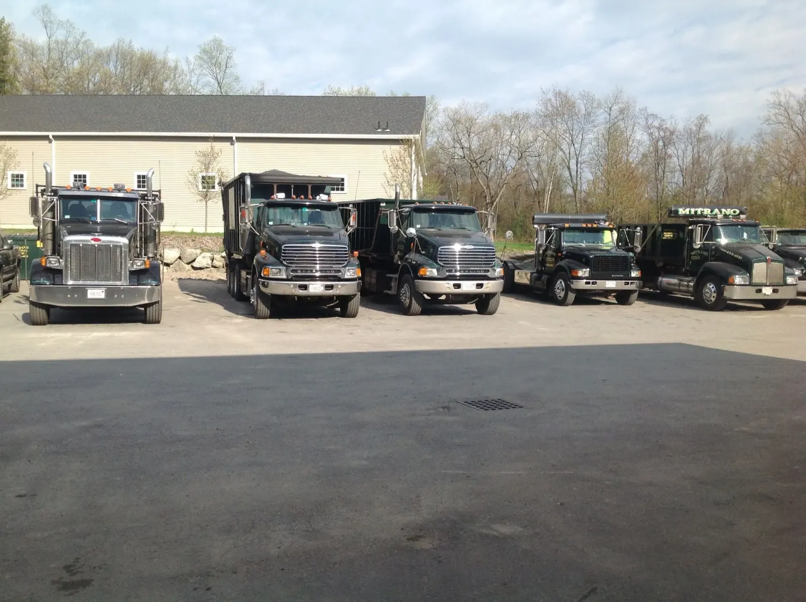 Five dark semi-trucks parked in a lot in front of a building on a sunny day.