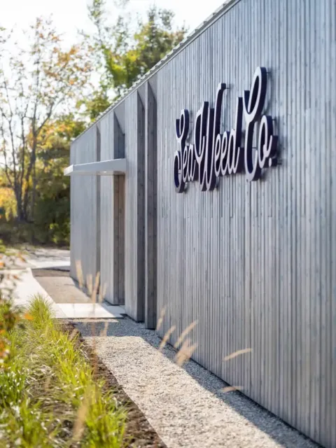 A modern wood-paneled building designed by a Maine architect, with Sea Weed Co. in large cursive letters on the exterior, surrounded by greenery and trees.