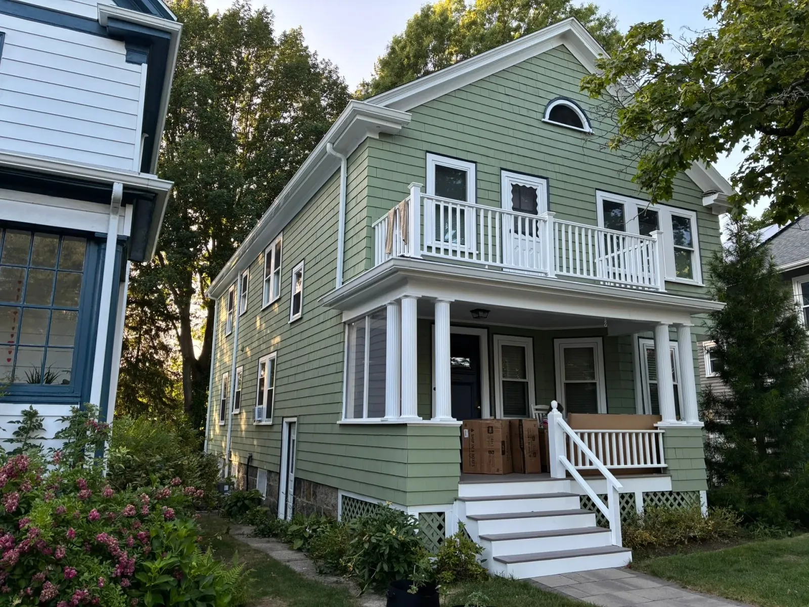 Green two-story house with white trim, front porch, and balcony on a tree-lined street