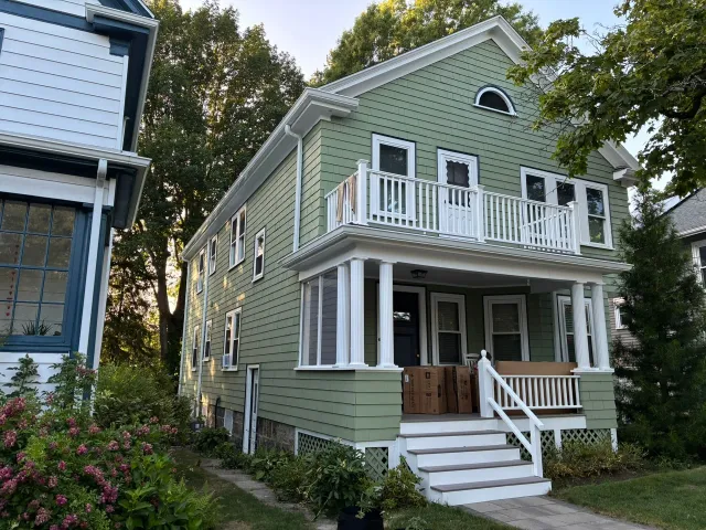 Green two-story house with white trim, front porch, and balcony on a tree-lined street