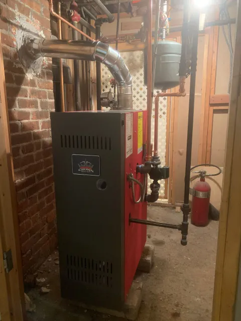 A red boiler is sitting in a basement next to a fire extinguisher.