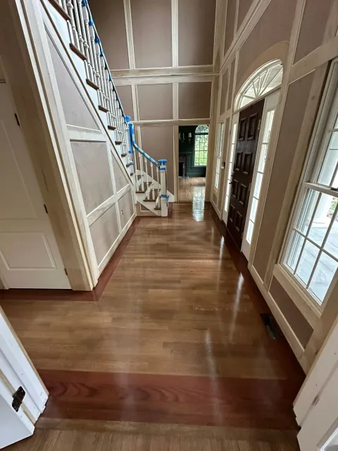 Hallway with hardwood floor, staircase, front door, and windows. Walls are beige with white trim.