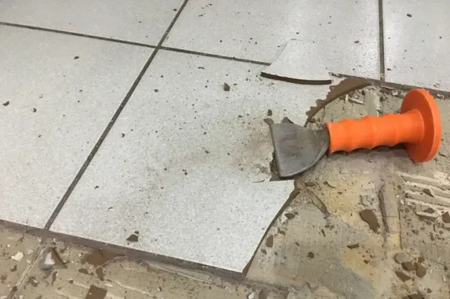 Broken floor tile with debris and an orange hand tool on a dusty tiled floor.