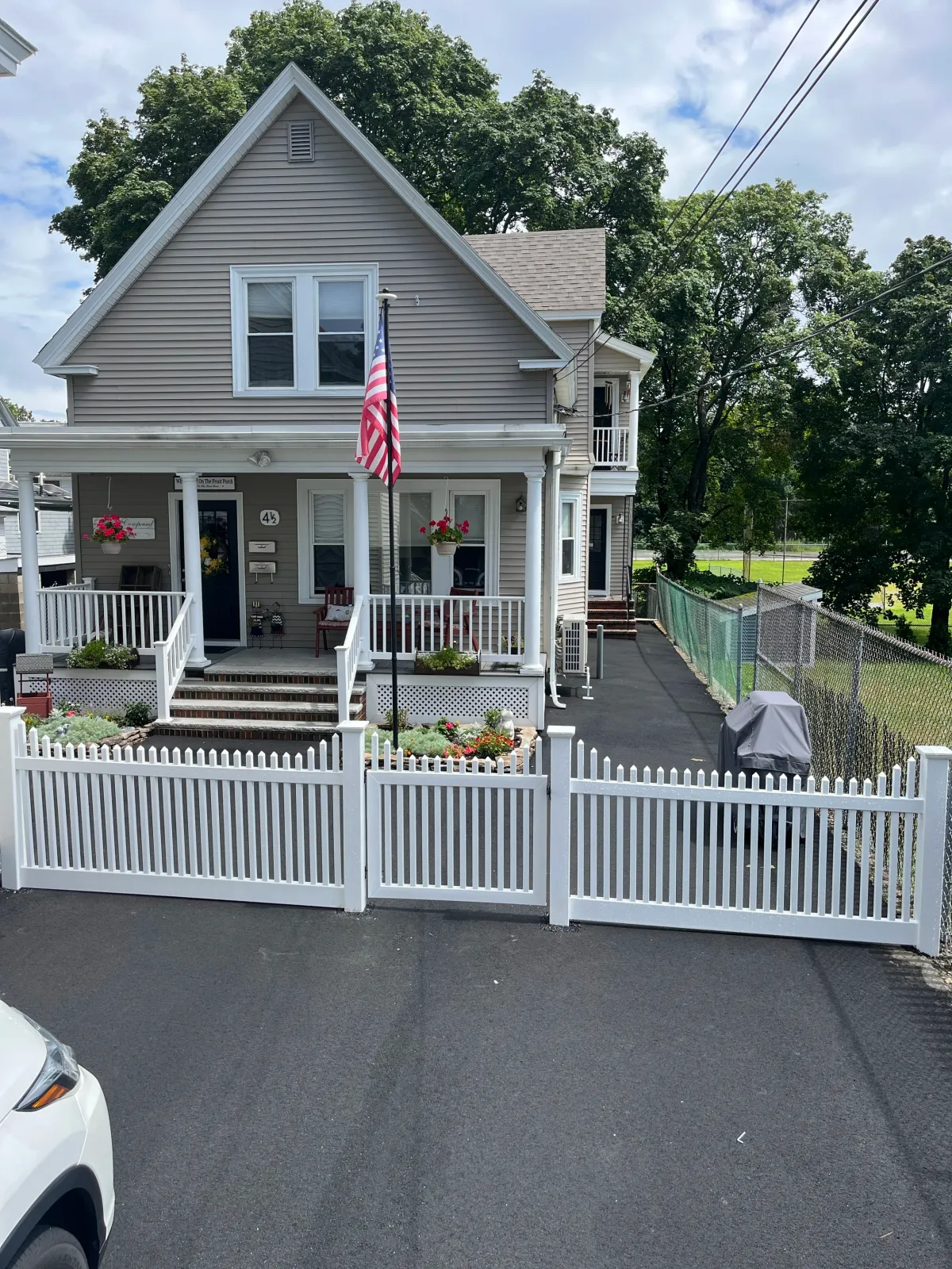 A light gray two-story house with front porch, white picket fence, American flag on pole, and flower pots. The driveway is visible in the foreground, and a parked white vehicle is partially in the frame. The house has multiple windows, a balcony on t