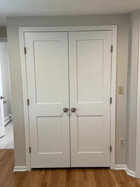 White double door closet with round silver knobs, against a wall painted light gray, with a baseboard heater and electrical outlets nearby, on a wooden floor.