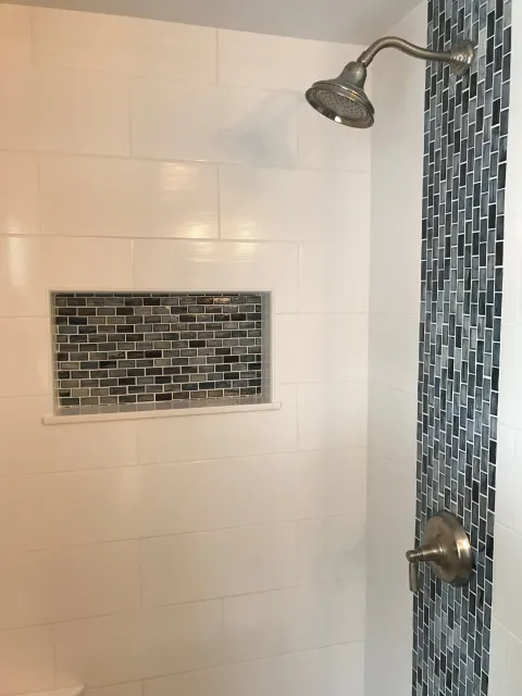 Bathroom shower with large white tiles on the wall, a small horizontal window with dark gray mosaic tiles, a chrome showerhead, and a matching chrome water control knob.