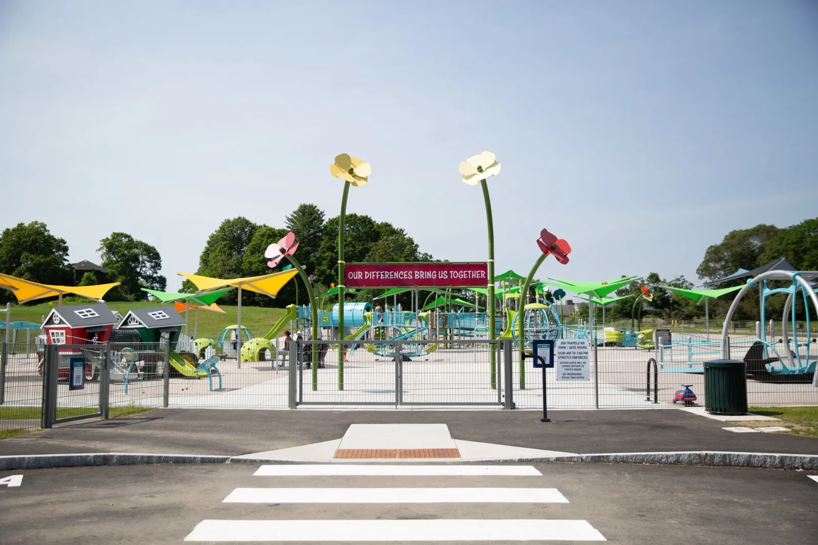 Colorful playground with slides, swings, and climbing structures, surrounded by a fence. A sign reads 'Our Differences Bring Us Together.' Crosswalk in foreground, blue sky overhead.