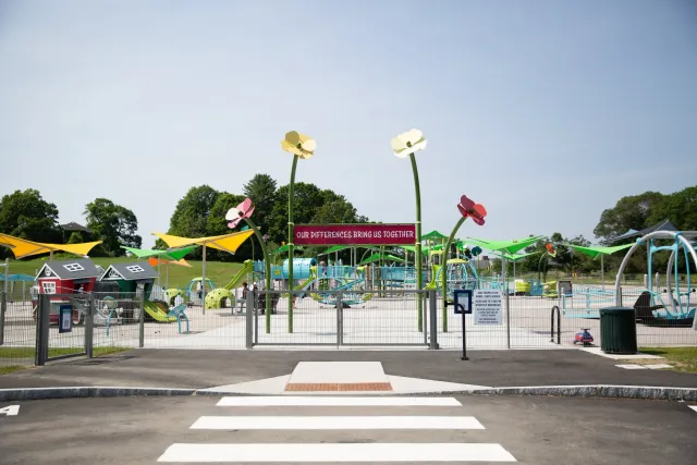 Colorful playground with slides, swings, and climbing structures, surrounded by a fence. A sign reads 'Our Differences Bring Us Together.' Crosswalk in foreground, blue sky overhead.