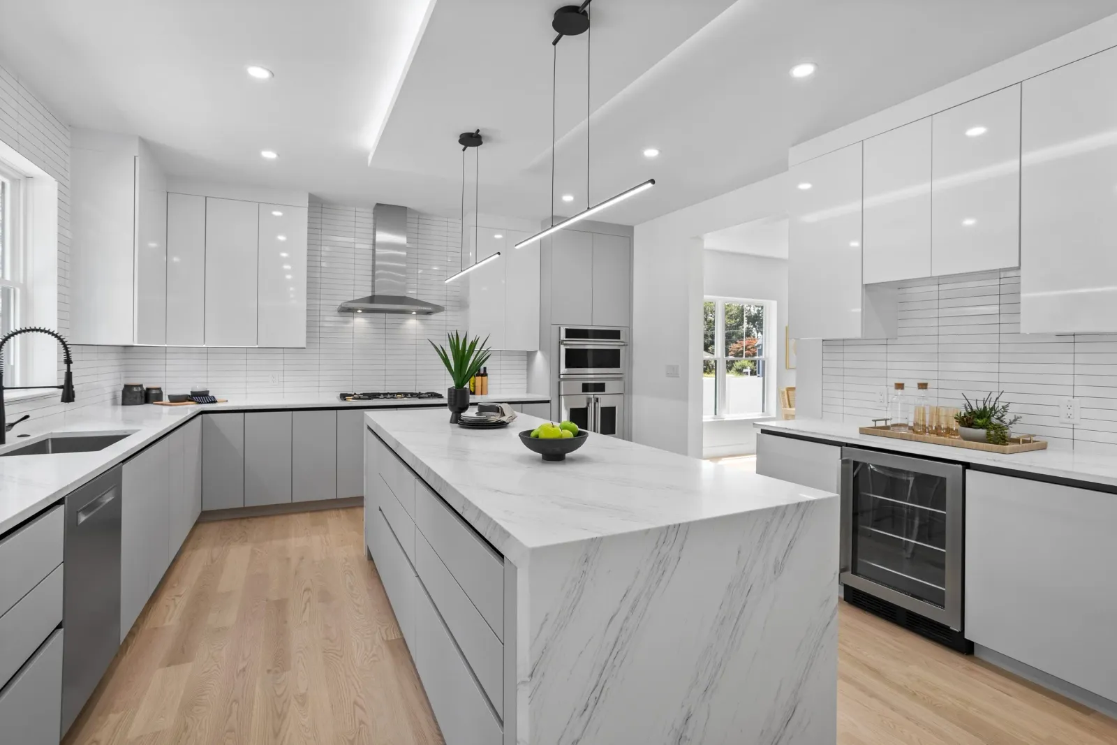 Modern white kitchen with marble island, stainless steel appliances, and light wood flooring, bright natural lighting from windows.