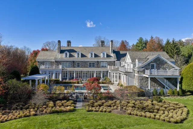Large stone and white siding house with multiple chimneys, surrounded by landscaped gardens and a pool in the backyard under a clear blue sky.