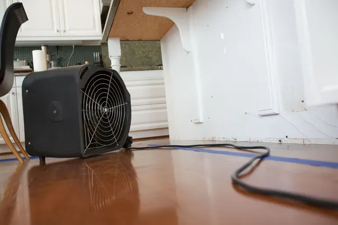 Black portable heater on a wooden floor beside a white wall, with a cord in the foreground