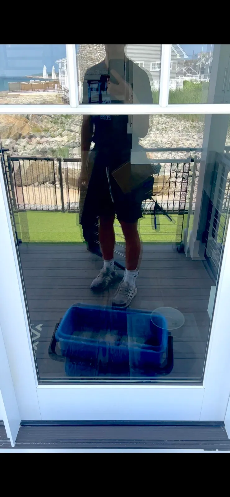 Reflection of a man taking a photo with a phone in a glass door during daytime, with his surroundings visible behind him, including a yard and neighboring houses.