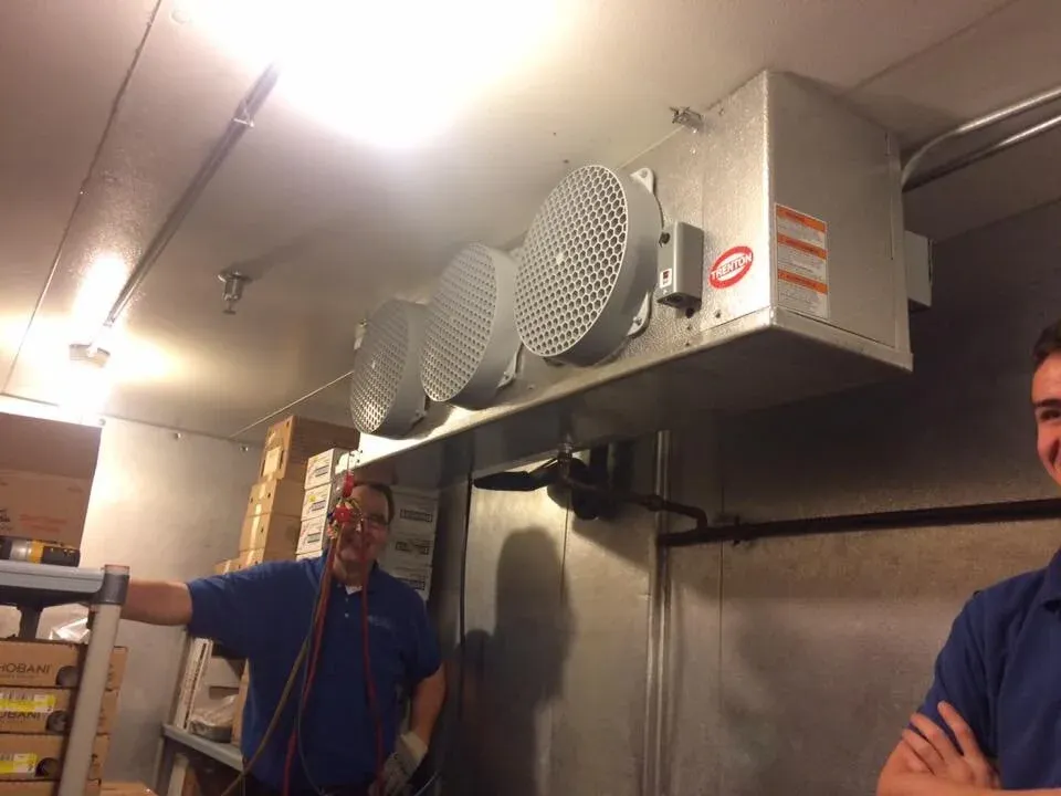 Two men in a walk-in refrigerator with cooling units and stacked boxes.