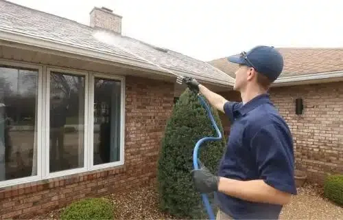 Man power washing a home's roof, performing exterior cleaning with a high-pressure hose. He is wearing a blue cap and gray shirt.