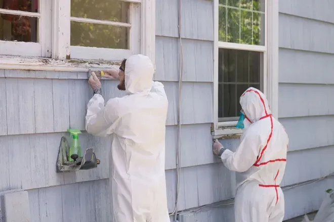 Two workers in white protective suits painting the exterior wall of a house