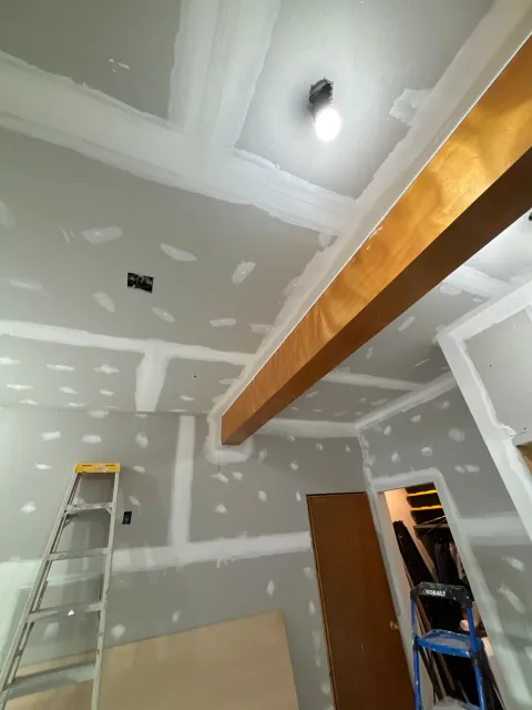 Interior room under construction with drywall, electrical outlets, a closed wooden door, stepladders, and a large wooden beam on the ceiling.