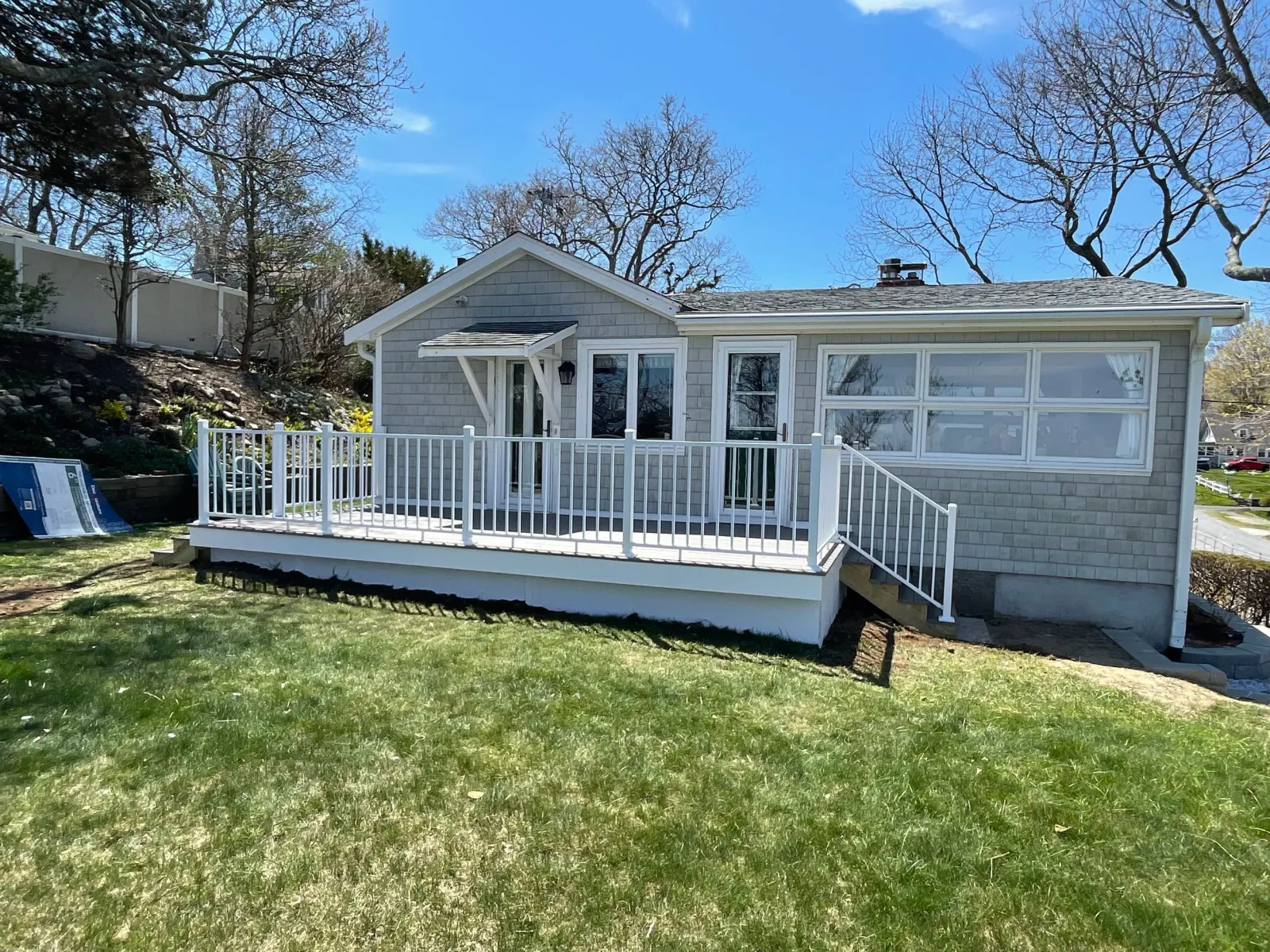 A single-story house with a small elevated porch with white railing, large front window, gray exterior siding, and a sloped roof. There are stairs leading up to the porch from the grassy backyard. Several trees are visible in the background and sky i