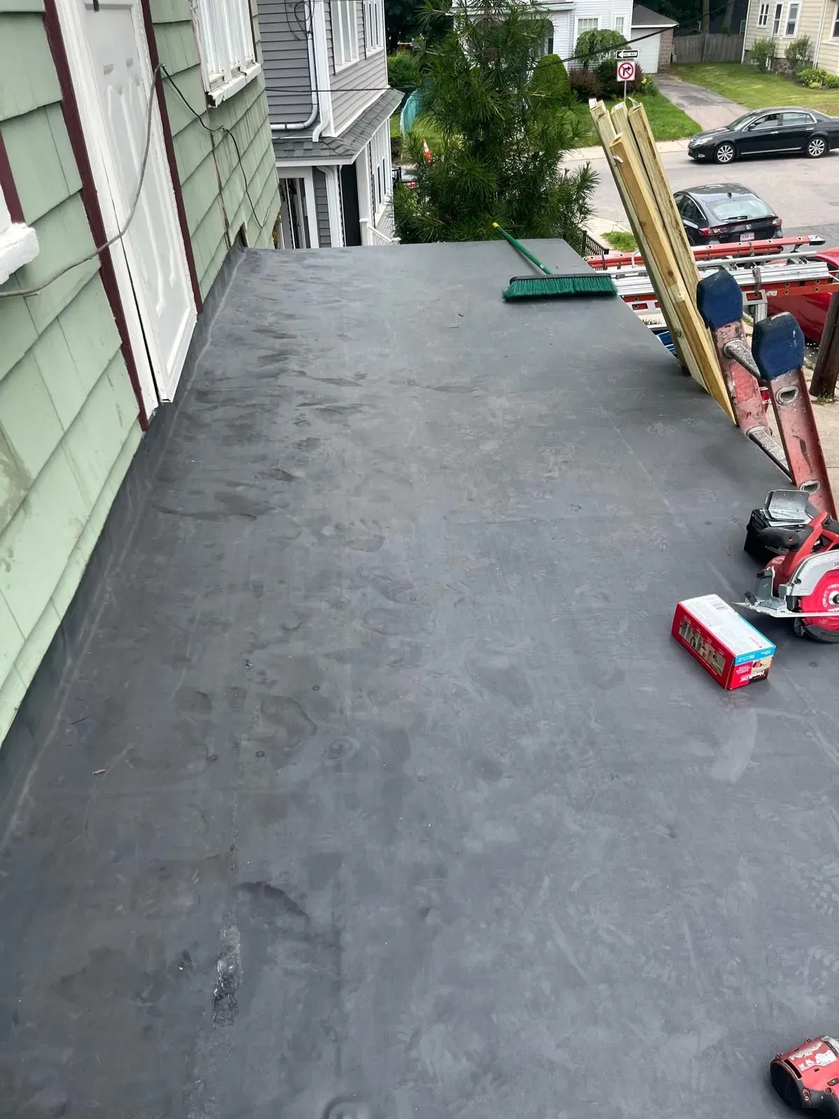 Flat rooftop with gray waterproofing beside a house, overlooking a street and parked cars below