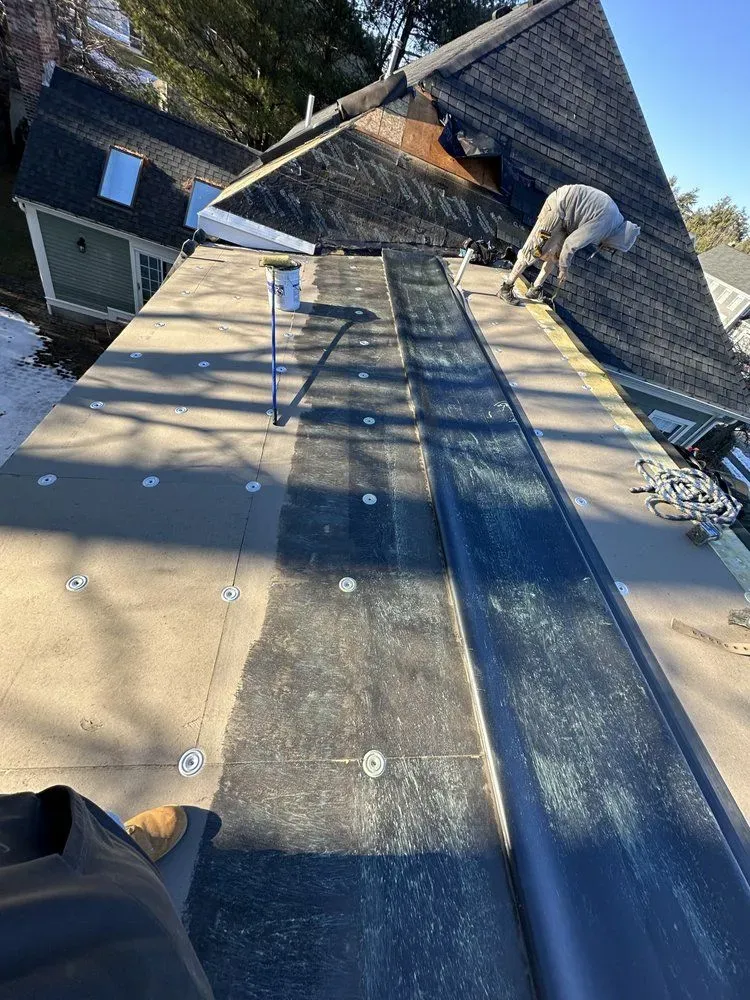 Worker repairing roof near chimney on a steep shingled rooftop with safety gear visible