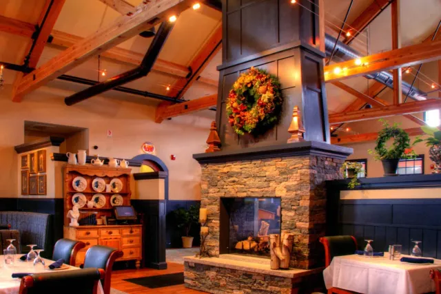 Interior of a cozy restaurant featuring a stone fireplace with a wreath, wooden beams on the ceiling with string lights, and wooden furniture including tables and chairs.