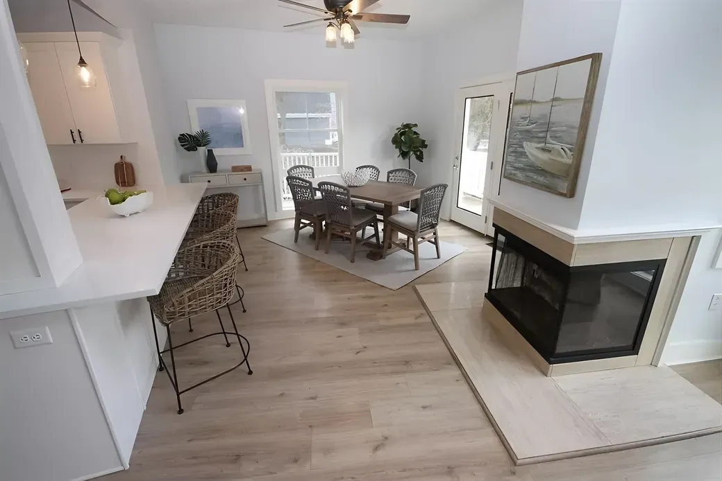 A bright, open-plan dining and kitchen area with a wooden table, woven chairs, a white breakfast bar, and a fireplace.