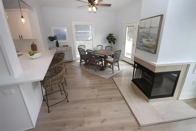 A bright, open-plan dining and kitchen area with a wooden table, woven chairs, a white breakfast bar, and a fireplace.
