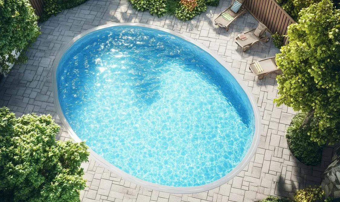 An oval swimming pool with turquoise water, surrounded by a light-colored paved patio and trees.