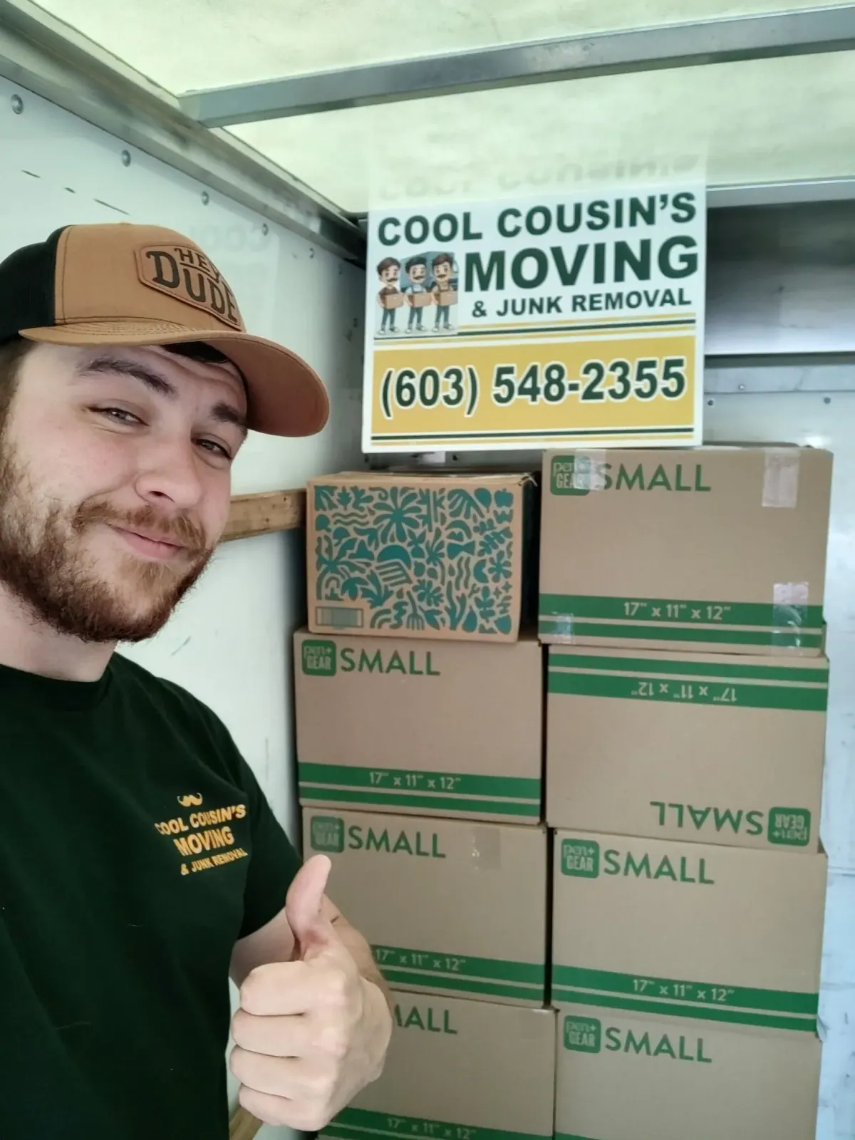 Man taking a selfie beside stacked moving boxes and a sign for Cool Cousin’s Moving & Junk Removal