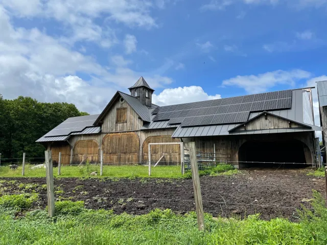 A rustic barn with a metal roof and solar panels, surrounded by green grass and a wire fence, under a blue sky with clouds and trees in the background.