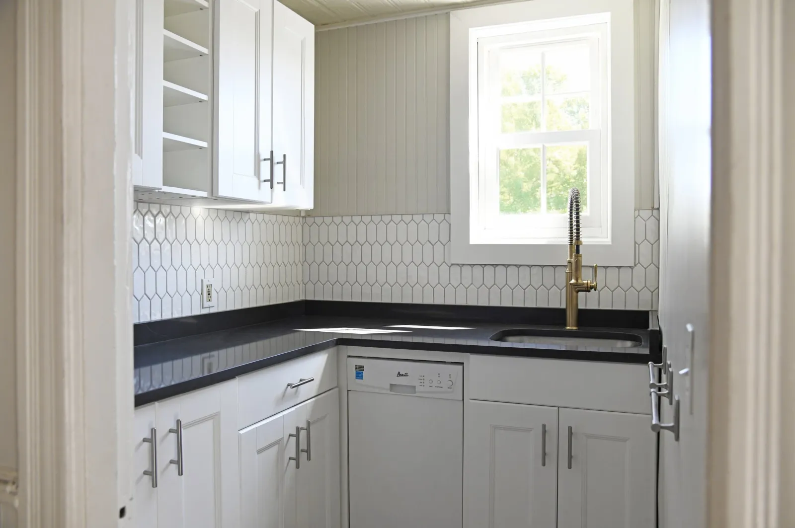 A small kitchen with white cabinets, a black countertop, an open shelf, a window, a gold faucet, and a white dishwasher.