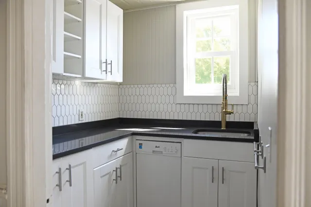 A small kitchen with white cabinets, a black countertop, an open shelf, a window, a gold faucet, and a white dishwasher.