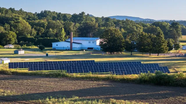 A rural scene with solar panels installed on land, a white building with a red chimney, trees, and hills in the background.