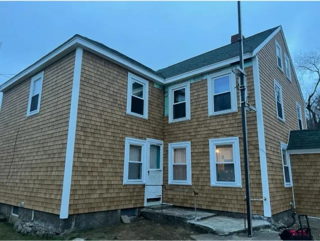 Rear view of a tan vinyl-sided house with white trim and a brick foundation under a blue sky